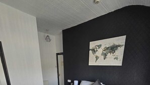 A neatly made bed with white linens and two pillows, a world map on the wall, and a small bedside table with a lamp.