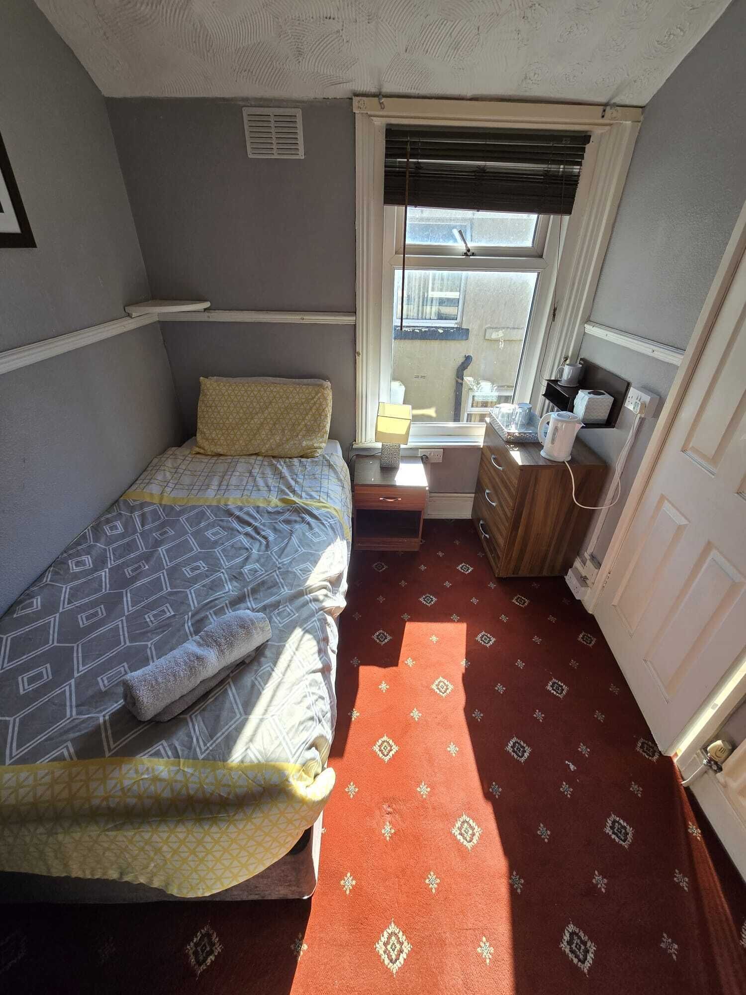 A small, compact room with a bed, a nightstand, a window with blinds, and a radiator.