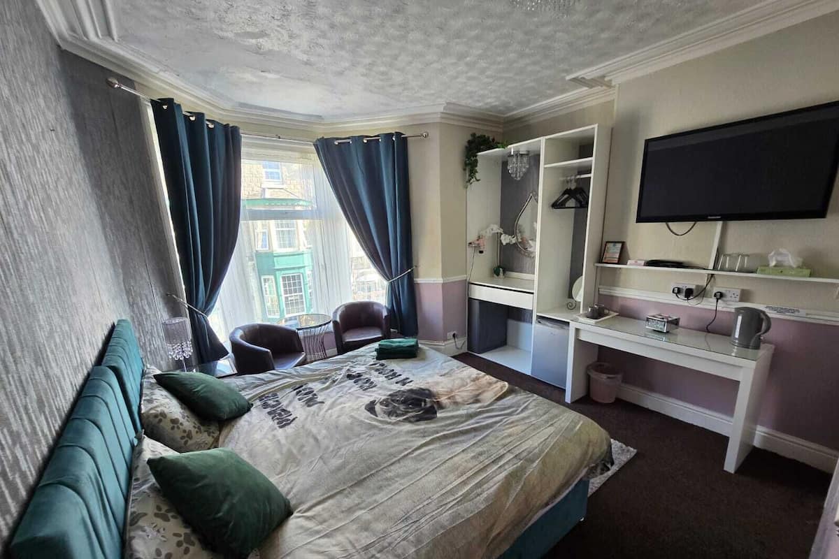 A bedroom with a bed, a desk, a chair, a television, and a window with curtains.