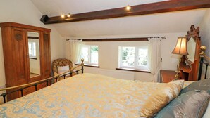A bedroom with a large bed, a wooden wardrobe, a chair, a window with curtains, and a bedside table with a lamp.
