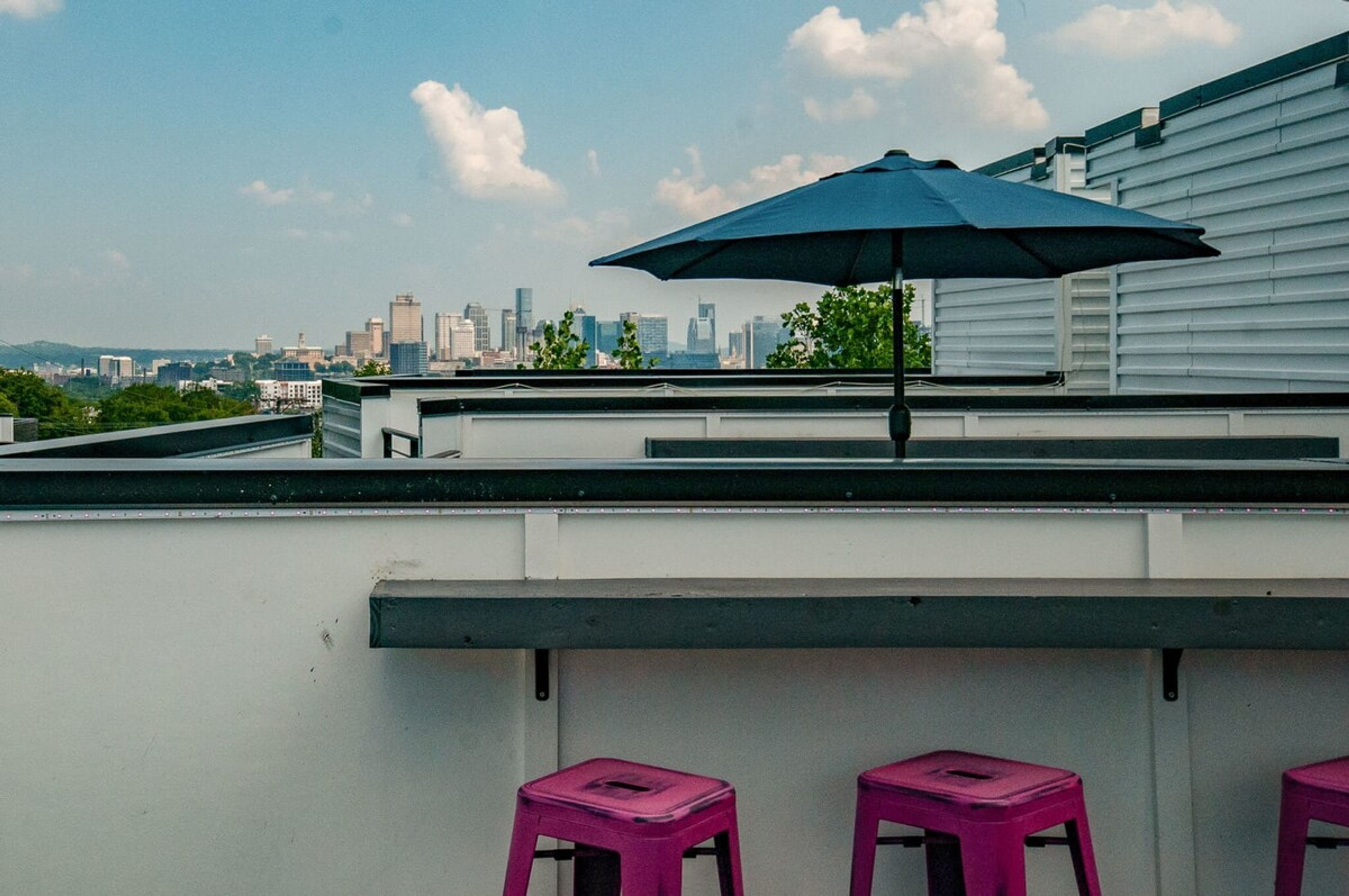 En tagterrasse med udsigt over byens skyline, en blå parasol og to pink barstole.