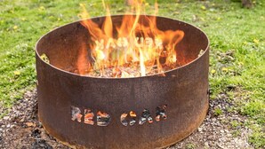 A metal fire pit with visible char marks, set on grass with a wooden deck and a house in the background. - Vacation Home Rental PM 51 (Russell)