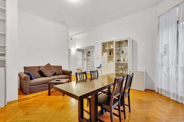 A modern living room with a wooden floor, a brown sofa, a dining table with chairs, and a white shelving unit.