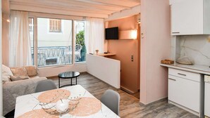 Een moderne woonkamer met een bank, eettafel en stoelen. Er is een balkon met uitzicht op groen en een gebouw. Een keuken met kastjes en een televisie is zichtbaar.