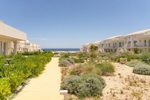 A pathway leading to a beachfront hotel with multiple-story buildings, a sandy area with shrubs, and a clear view of the sea.