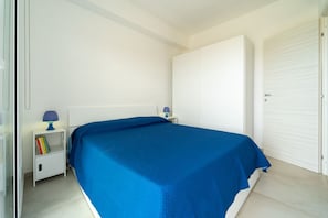 A bedroom with a bed, a nightstand with a lamp, a closed door, and a white wardrobe. - GuestHost - Charming 75 sqm seaview apartment on two levels, for 5 people, located inside the renowned Borgo 84 residence, equipped with swimming pool and cycle path. The property is located just 3 km from the center of Marzamemi, one of the most fascina (Pachino)