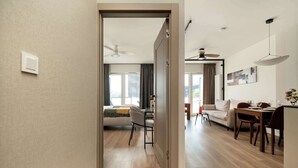 A modern hotel room with a wooden floor, a bed with a green throw, a dining area with a table and chairs, and a ceiling fan.