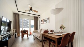 A modern living room with a dining area, a flat-screen TV, a ceiling fan, and a large window with curtains.