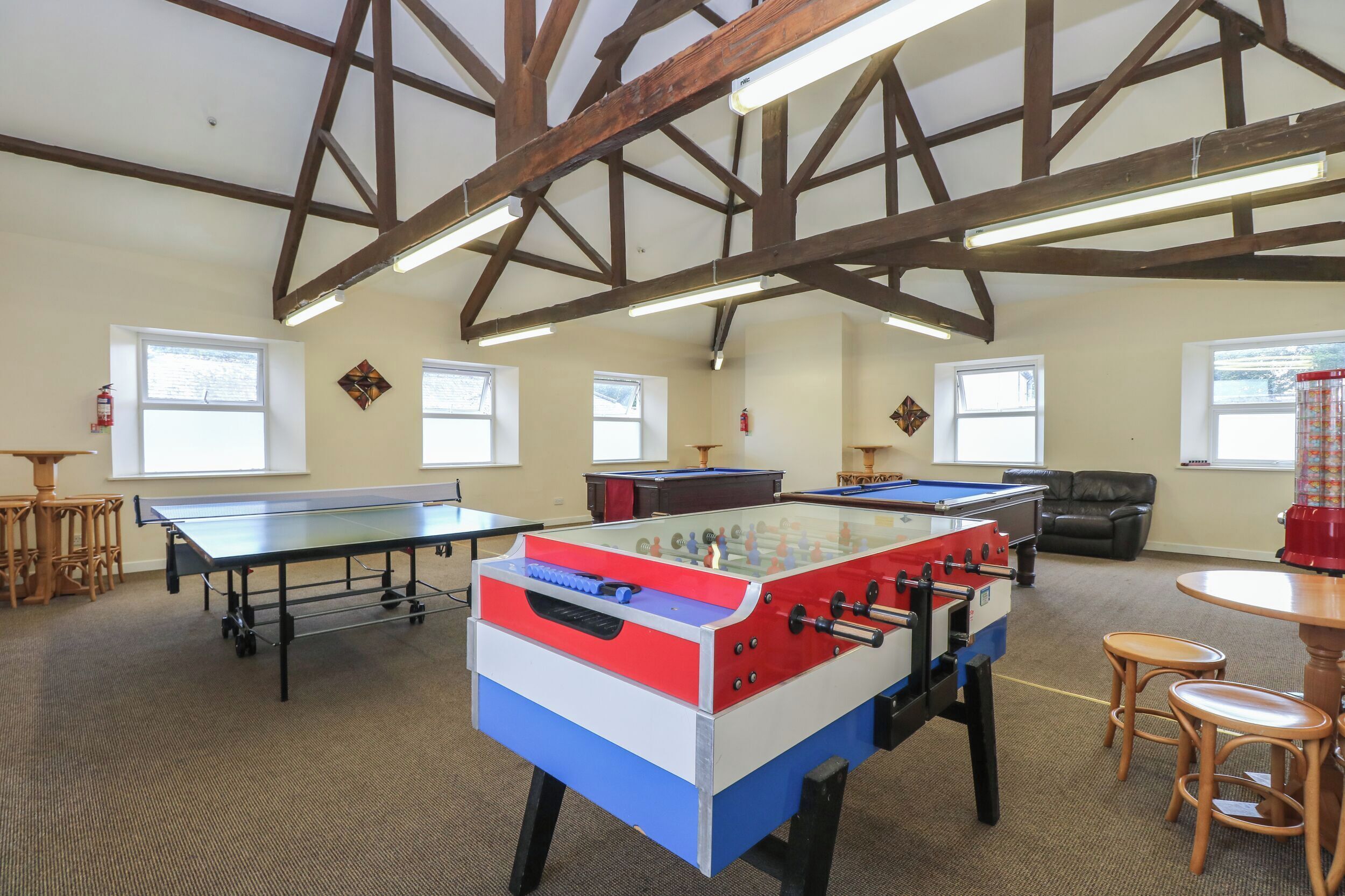 A room with a foosball table, ping pong table, and a pool table. There are windows, a fire extinguisher, and a leather chair.
