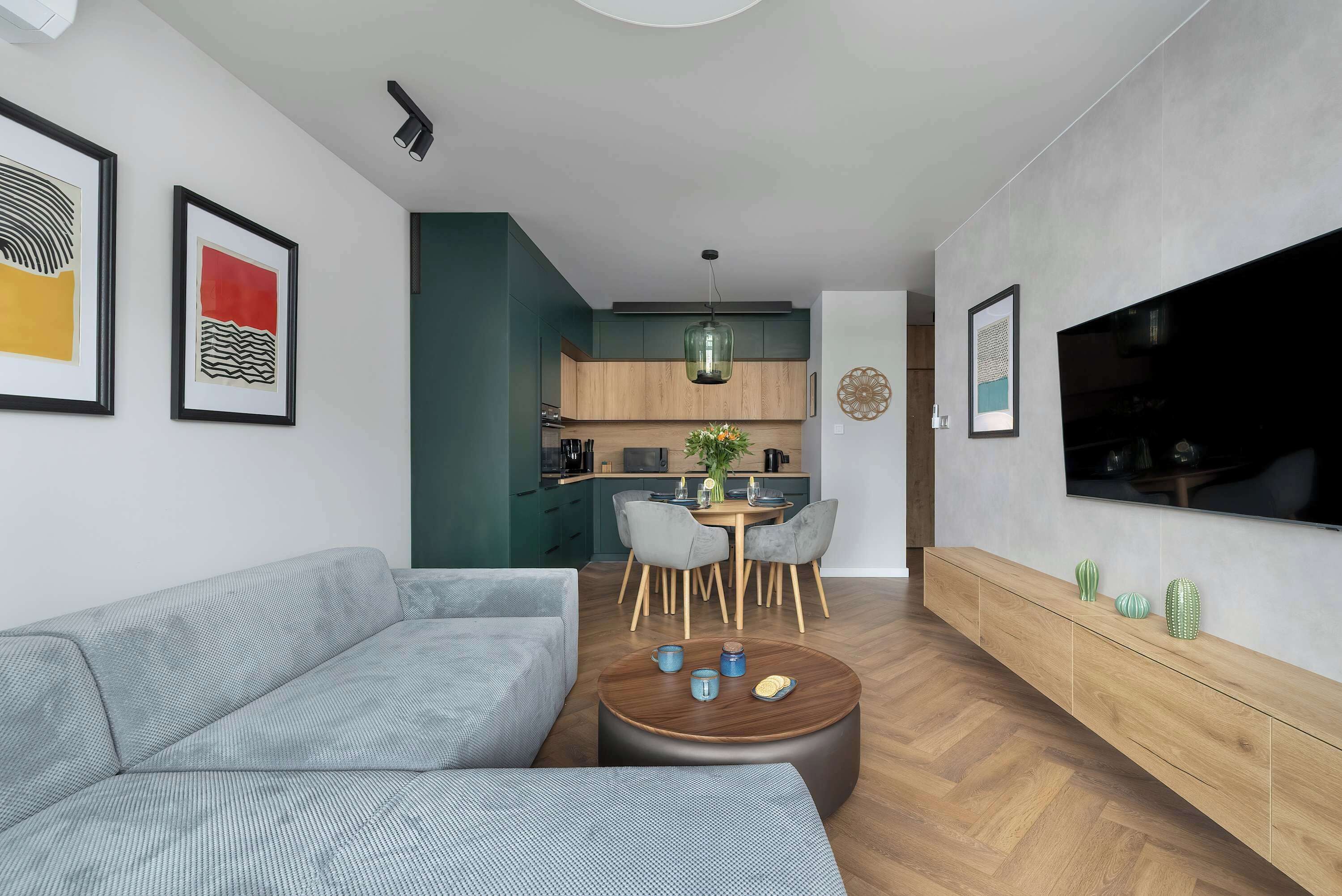 A modern living and dining area with a gray sofa, wooden table, green accent wall, and wall art for a cozy atmosphere.