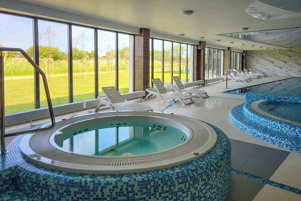 A modern indoor pool area with a hot tub, lounge chairs, and large windows offering a view of the outdoors.
