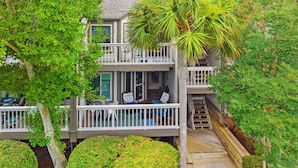 Een tweelaags huis met een balkon, omgeven door weelderige begroeiing en een palmboom.