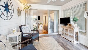 Een knus woongedeelte met een plafondventilator, een groot raam met zonwering, een televisie en een rieten fauteuil.