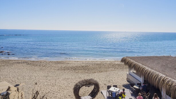 Un restaurante en la playa con terraza, techo de paja y vistas al mar.