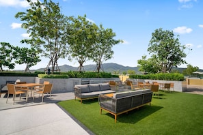 An outdoor patio with a dining set, sofa, and chairs, surrounded by greenery and trees, with a view of distant hills.