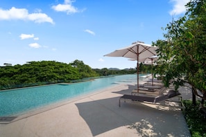 A swimming pool with lounge chairs and umbrellas, surrounded by greenery and a clear sky.