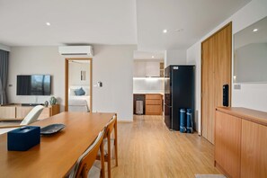A modern hotel room with a wooden dining table, a flat-screen TV, a bed, and a kitchen area with a refrigerator and cabinets. - Reef Residences, Three-Bedroom 3505 3B Premium (Choeng Thale)