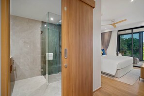 A modern bedroom with a glass shower enclosure, a wooden door, a large bed, and a view of greenery through the window.