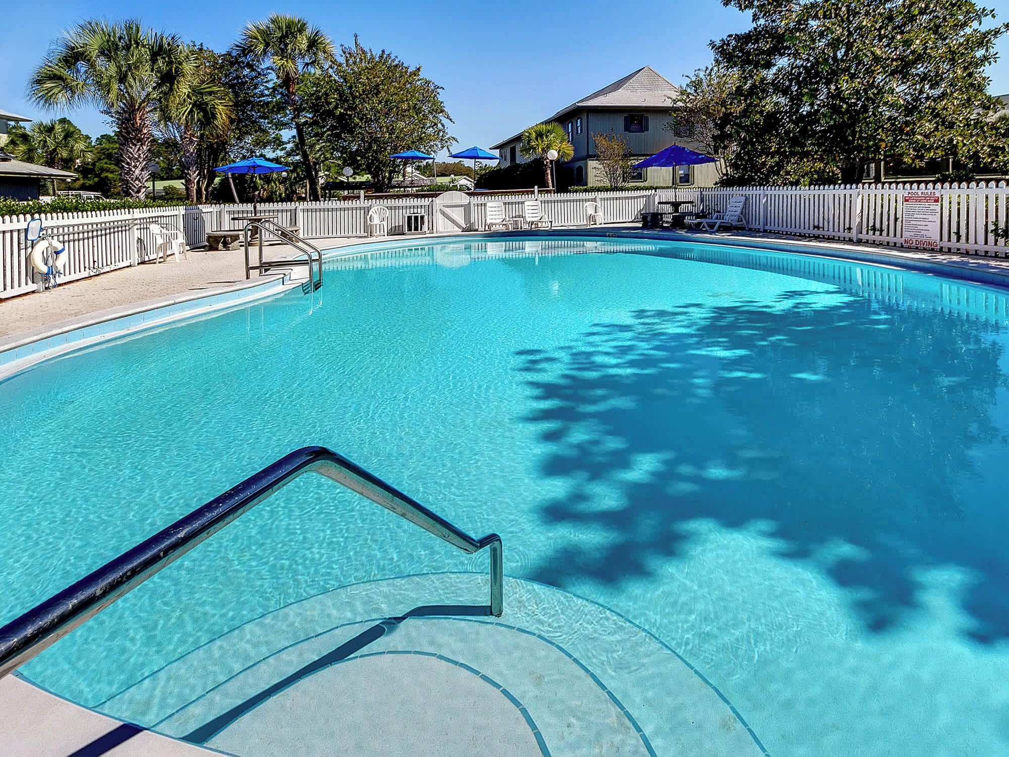 A clear swimming pool with a white fence, blue umbrellas, and a sign indicating pool rules.
