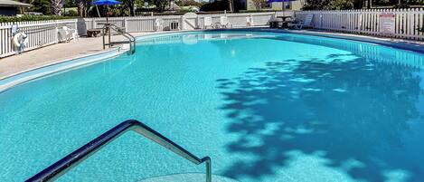 A clear swimming pool with a white fence, blue umbrellas, and a sign indicating pool rules.