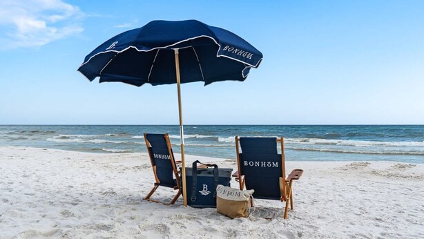 Une scène de plage avec deux chaises de plage en bois, un parasol bleu et un sac de plage.