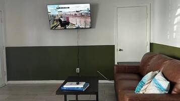 Een woonkamer met een aan de muur bevestigde televisie, een bruine leren bank en een klein zwart salontafeltje met een boek.