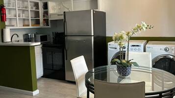 A compact kitchen with a refrigerator, microwave, and washing machine. There is a dining table with chairs and a vase of flowers.