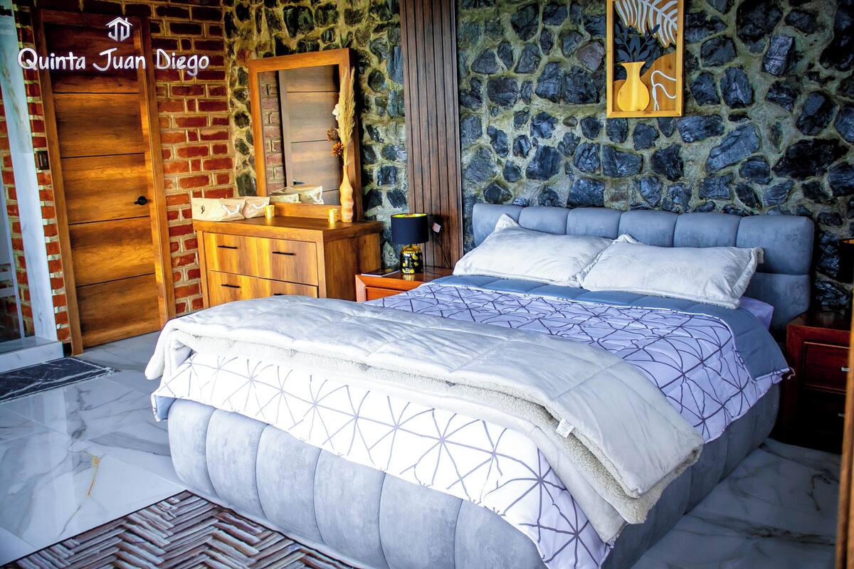 A bedroom with a large bed, a wooden dresser, a stone wall, and a mirror.