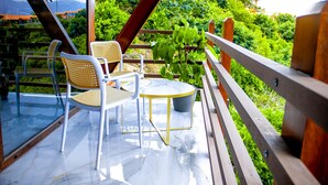 Un espace extérieur aménagé avec une structure en verre, comprenant deux fauteuils en osier et une petite table ronde, le tout entouré d’une balustrade en bois et d’une végétation luxuriante.
