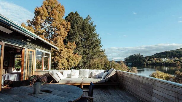 Una terraza de madera con una mesa redonda y sillas, un sofá con cojines, y vistas a un lago y árboles.