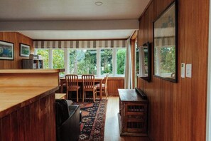 A cozy kitchen with wooden cabinetry, a dining area with chairs, and large windows offering a view of greenery.