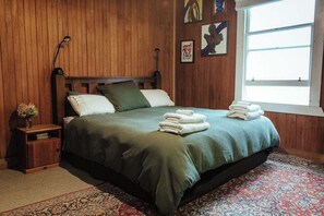 Un dormitorio con cabecera de madera, colcha verde, dos almohadas, mesita de noche con un florero y una ventana con vistas.