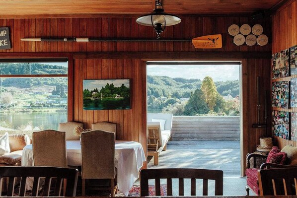 A cozy room with a wooden interior, a dining area, and a large window offering a scenic view of a lake and hills.