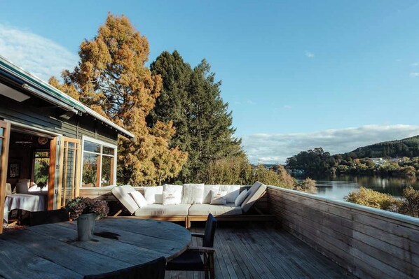 Una terraza de madera con una mesa redonda y sillas, un sofá con cojines, y vistas a un lago y árboles.