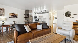 A modern living room with a leather sofa, a wooden coffee table, and a white armchair. A kitchen with white cabinets and a microwave is visible in the background.