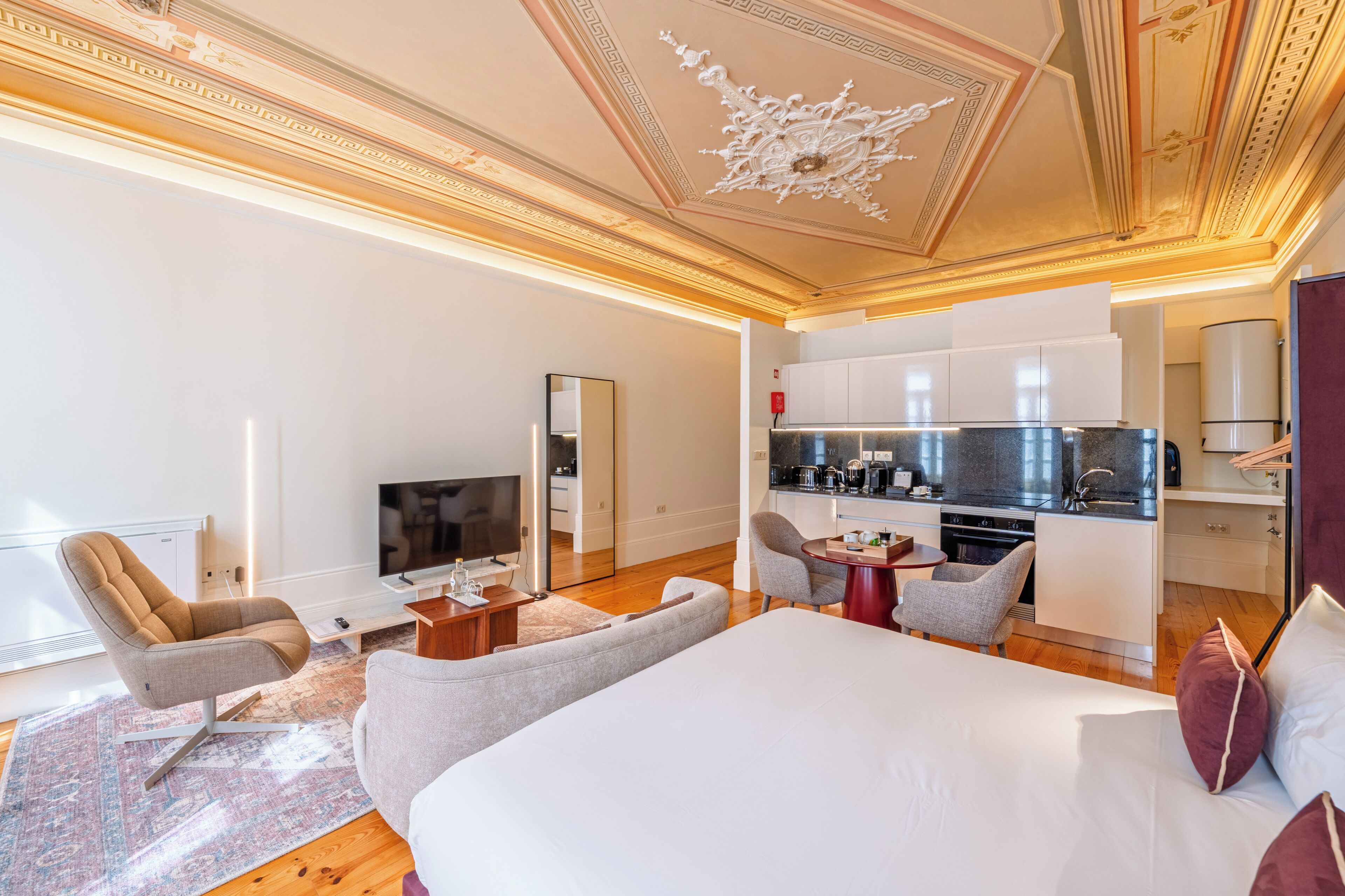 Una habitación de hotel moderna con una cama grande, una zona de estar con sofá y sillones, un televisor, una pequeña cocina y un techo decorativo.