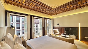 A hotel room with a large bed, a chair, a desk, a TV, and a balcony with a view of buildings. - YOUROPO - Collection (Porto)