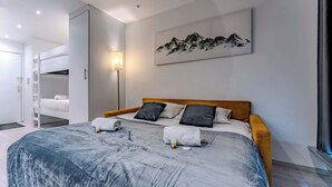 A modern bedroom with a bunk bed, a large bed with a grey and white duvet, a wooden headboard, and a mountain painting on the wall.