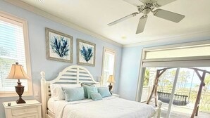 Un dormitorio con cama blanca, suelo de madera, ventilador de techo y vistas al exterior a través de un ventanal.