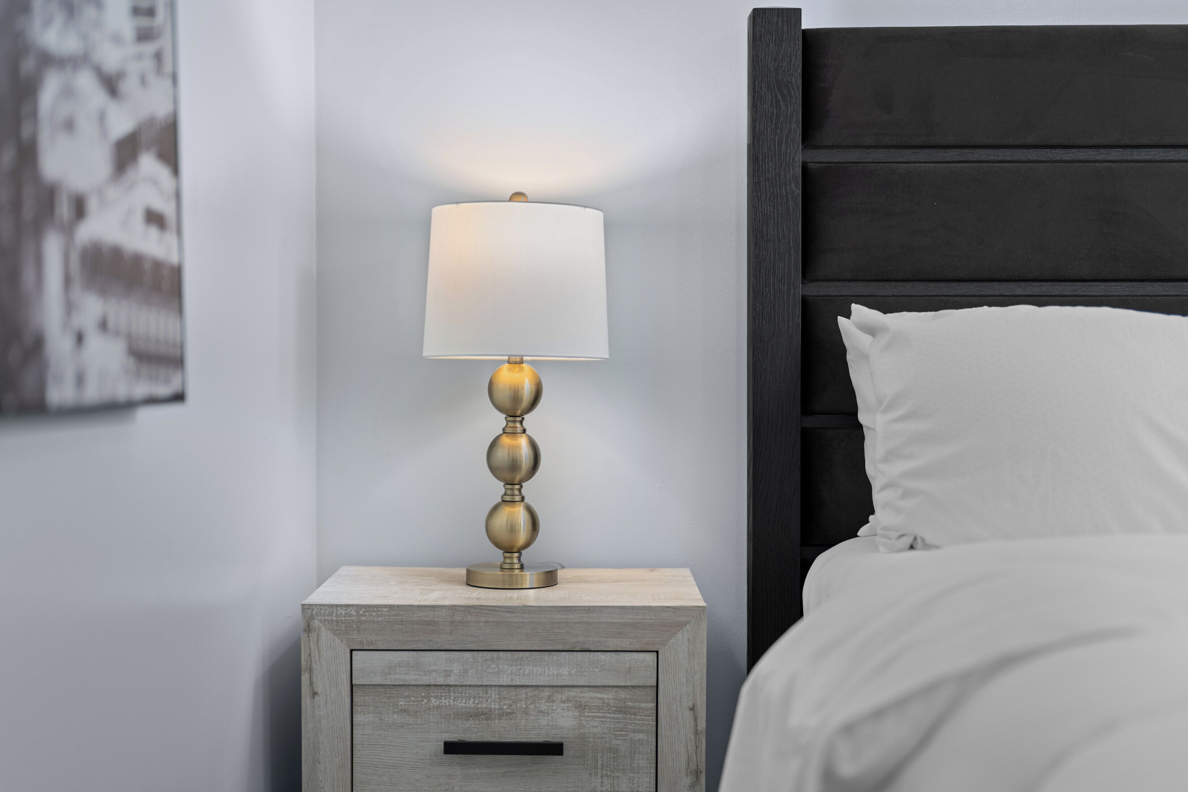 Une table de chevet avec une lampe, un tiroir, et un lit avec du linge de lit blanc.