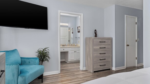 Un dormitorio con cama, sofá azul, cómoda, televisor y baño visible a través de una puerta abierta.