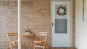 Un muro de ladrillo, dos sillas de madera, una mesa pequeña con un florero y una puerta con un adorno festivo.