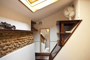 Uma escada estreita com uma parede de tijolos de um lado e uma prateleira de madeira do outro. Um ventilador está fixado na parede acima da prateleira.
