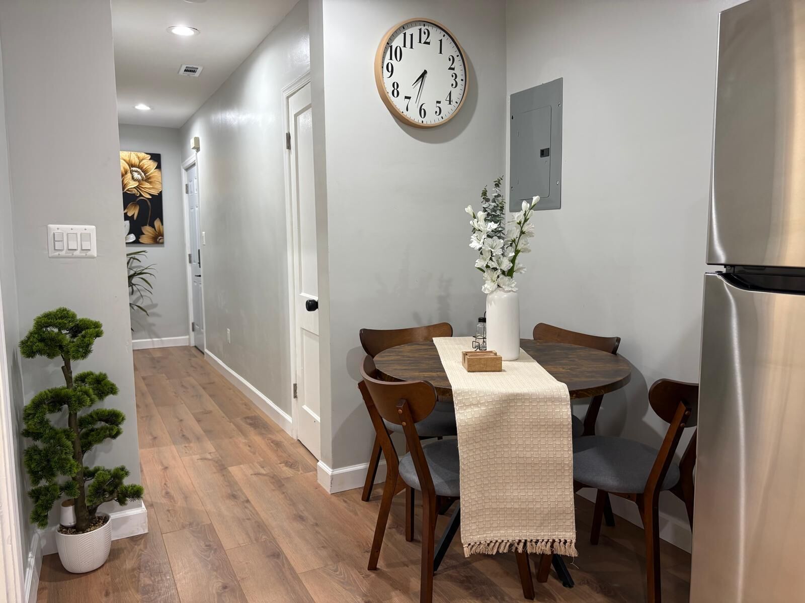 A modern kitchen with a round dining table, wooden chairs, a clock, and a refrigerator.