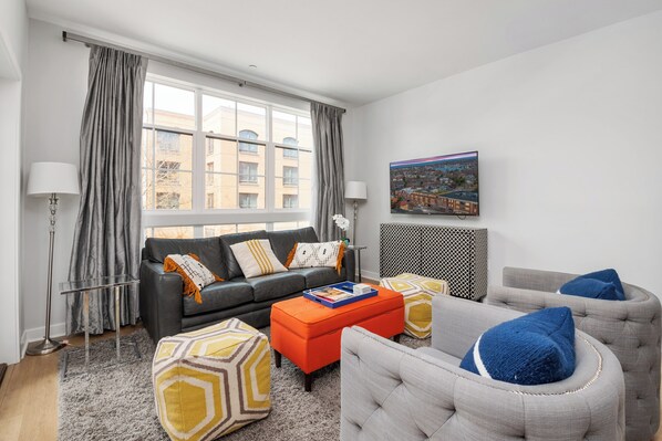 Ein modernes Wohnzimmer mit grauem Sofa, orangefarbenem Hocker und grauem Sessel. Ein großes Fenster mit Vorhängen und einem Stadtbild an der Wand.