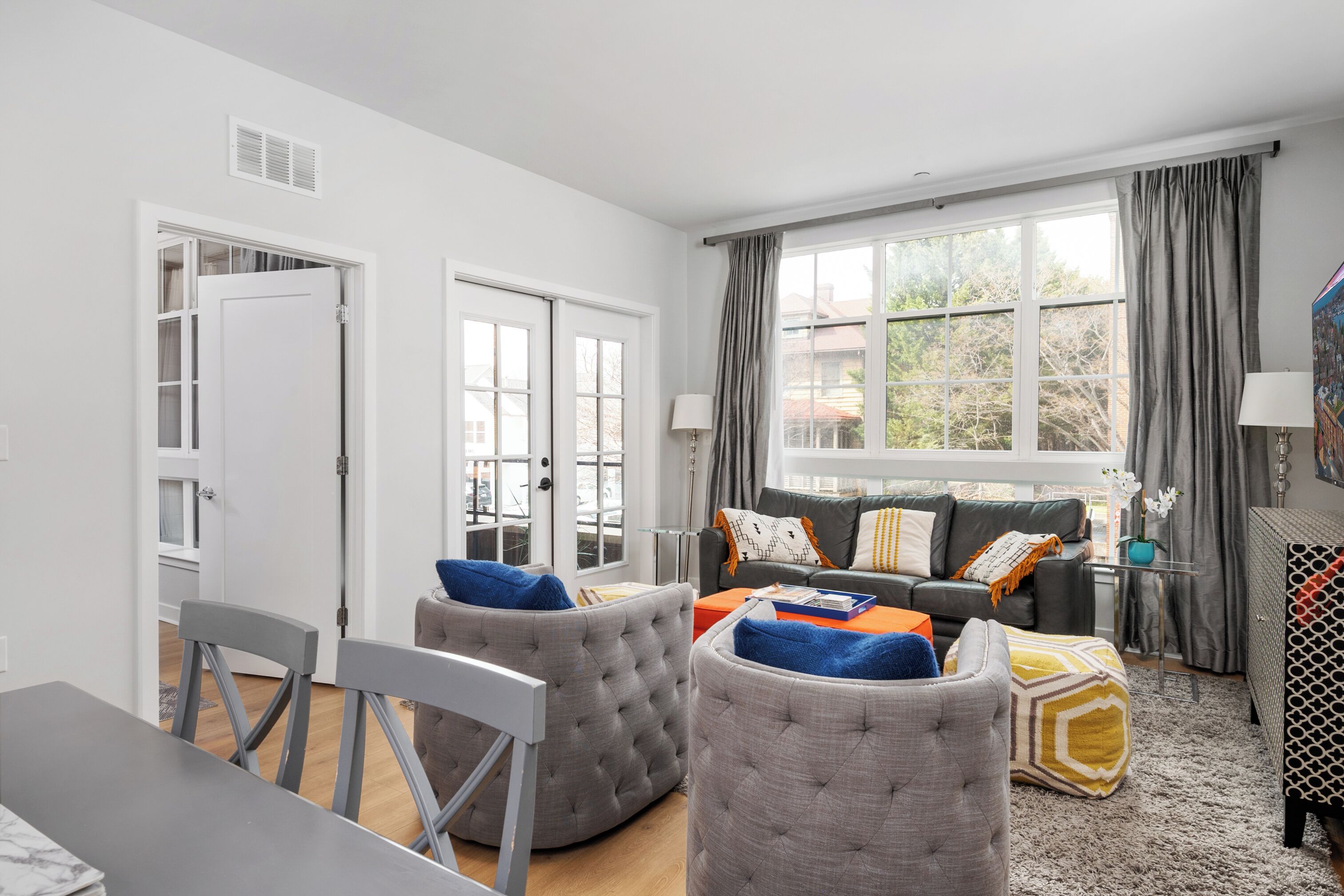 A living room with a grey sofa, ottomans, a dining table, and a window with curtains.