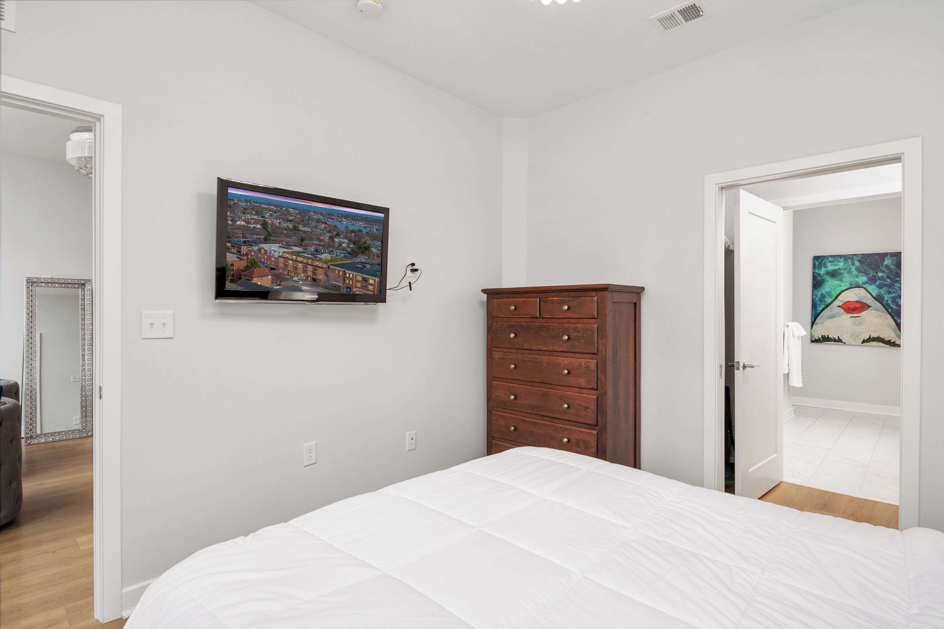 A bedroom with a bed, a dresser, a television on the wall, and a door leading to another room.