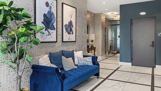 Un hall d’entrée moderne avec un canapé bleu, deux tableaux abstraits encadrés, une plante en pot et un sol en marbre.