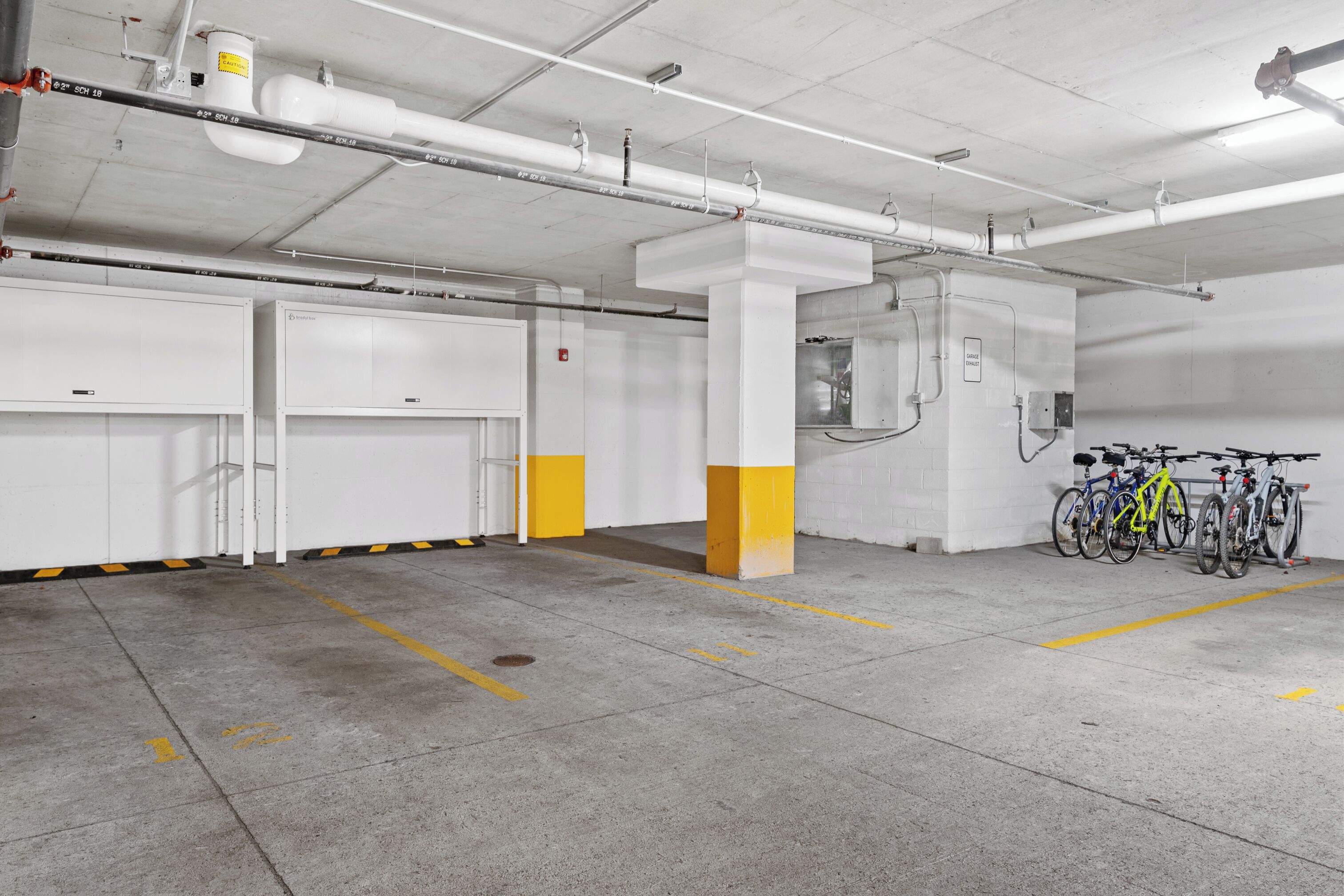 A clean, well-lit parking garage with concrete floors and white walls. There are several bicycles parked near the wall, and the garage has a modern industrial design with exposed pipes and a ventilation system on the ceiling.