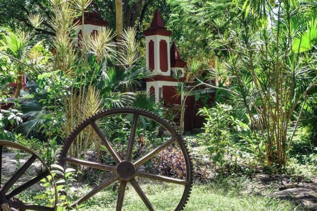 A garden with a large metal wheel, lush greenery, and a small red and white structure.